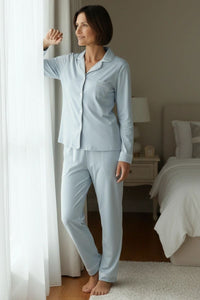 Woman in light blue pajamas standing by a window in a bedroom.